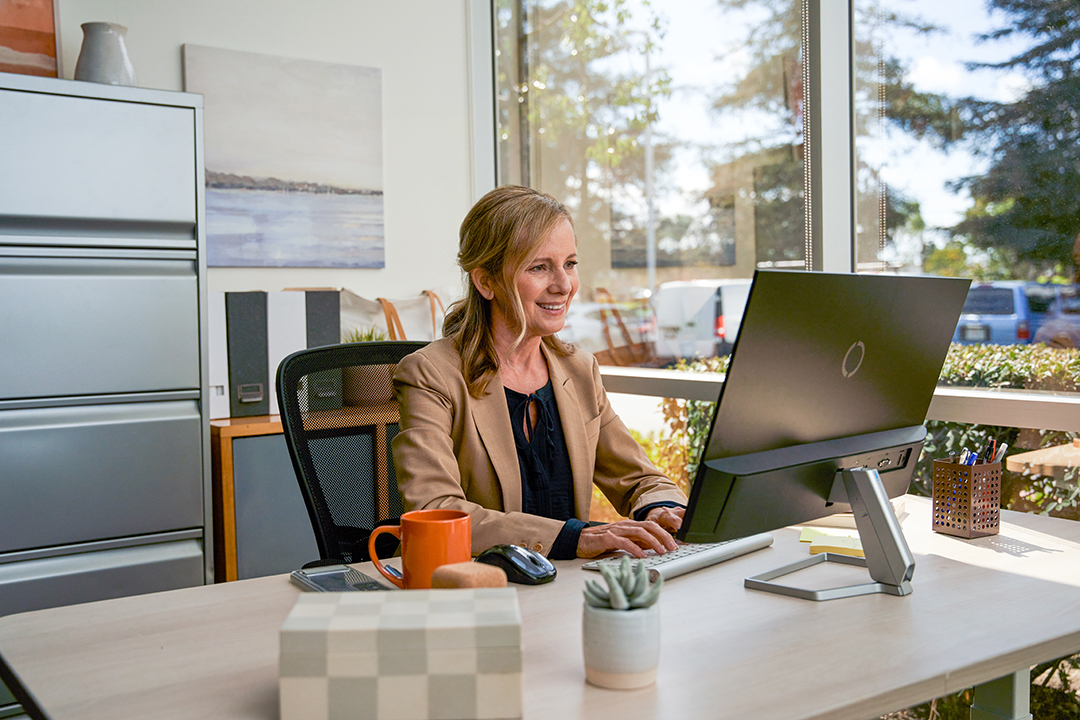 A professional woman sits at her desk in a bright office, smiling as she looks at her computer screen.